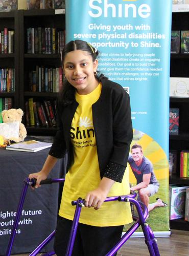 Girl with purple walker stands in a bookstore in front of Shine branded table set up