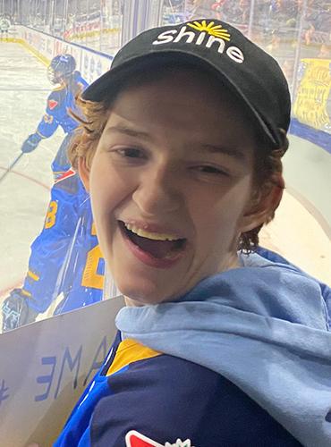 Young woman with curly hair looks at camera while standing behind the glass at a PWHL game, holding a cheer sign