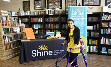 Teenage girl is in bookshop in front of a Shine Foundation display. She leans on her purple walker