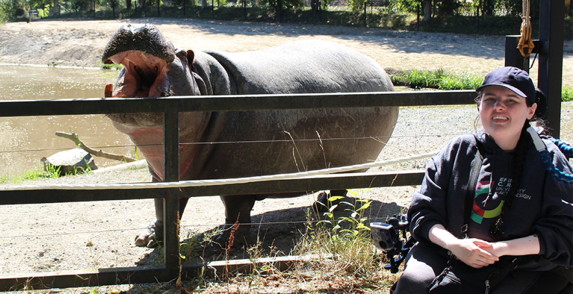 Young woman in power wheelchair near zoo habitat with an open-mouthed hippo