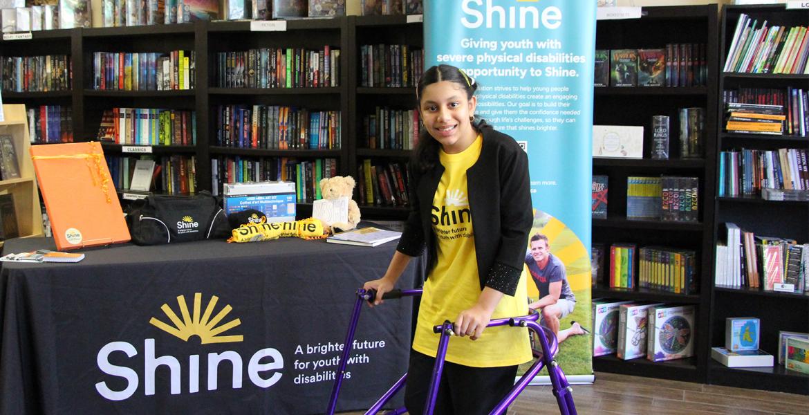 Girl with purple walker stands in a bookstore in front of Shine branded table set up