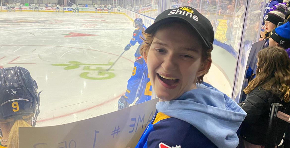 Young woman with curly hair looks at camera while standing behind the glass at a PWHL game, holding a cheer sign